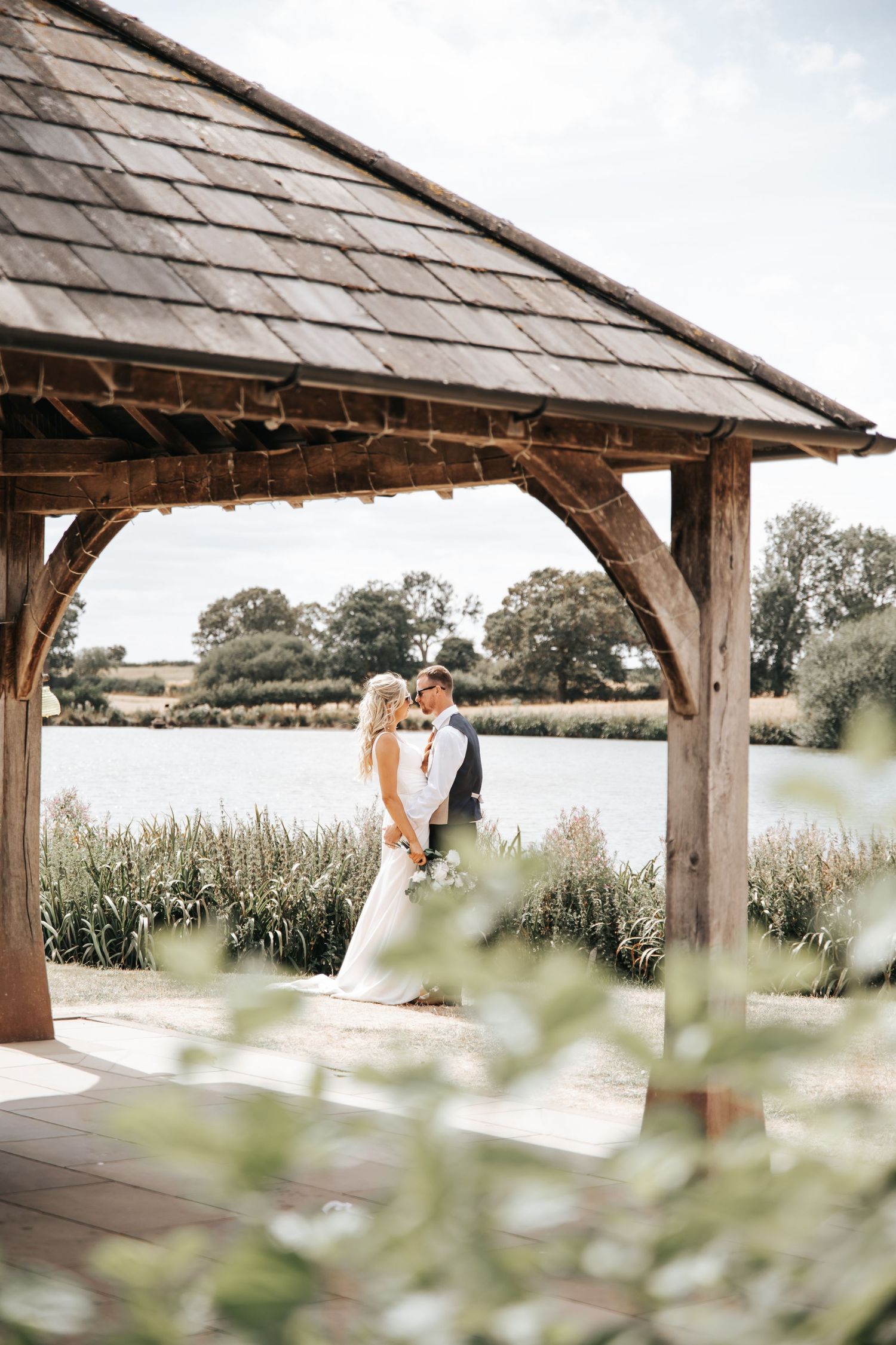 Bride and groom share a romantic moment under a rustic wooden gazebo beside a serene lake on their wedding day.