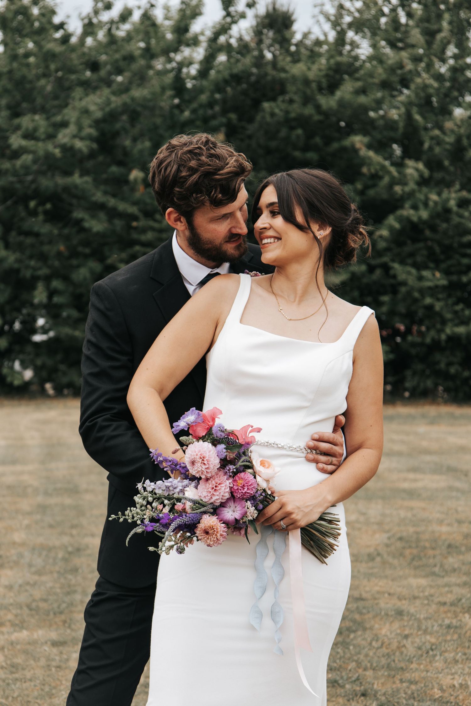 Wedding couple shares intimate moments outdoors with pink floral bouquet.