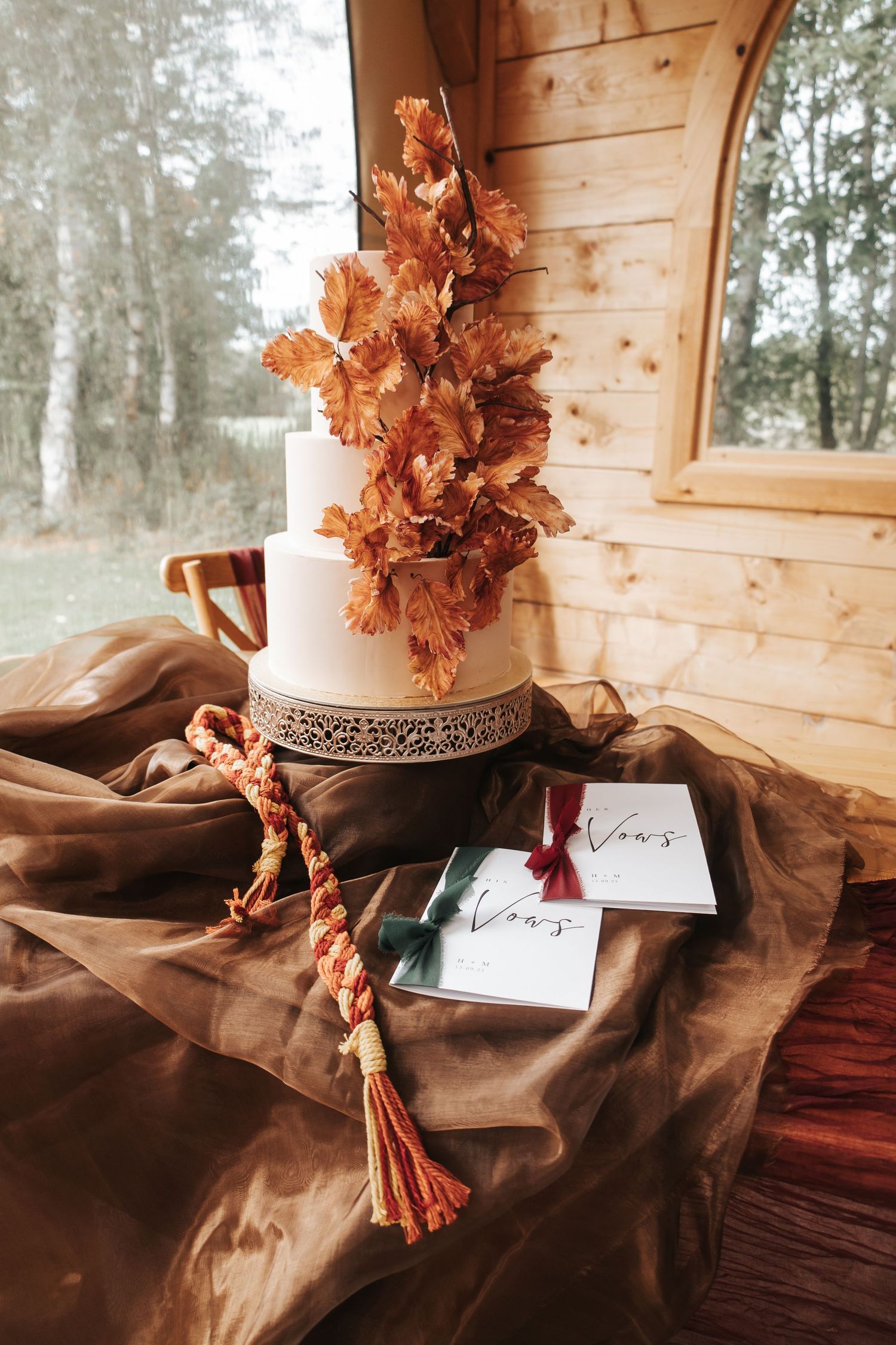 Wedding cake decorated with autumn leaves and brown fabric in rustic wooden setting.