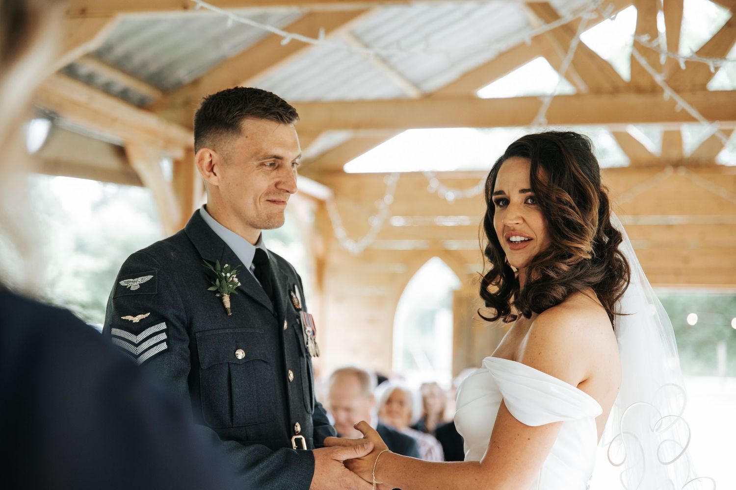 Military groom in uniform holds hands with bride in off-shoulder dress during outdoor wedding ceremony.