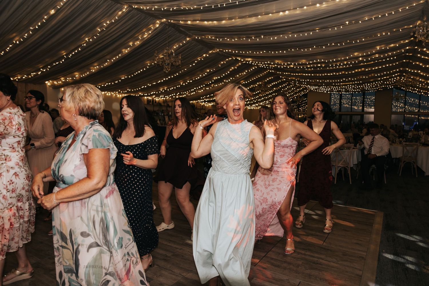 Women in elegant dresses dancing happily under fairy lights at a wedding reception.