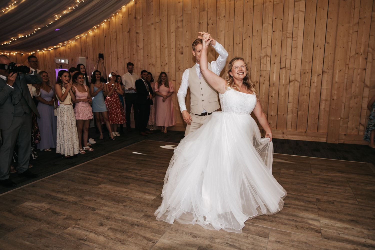 Bride and groom share their first dance in a warmly lit barn venue, surrounded by guests capturing the joyful moment.