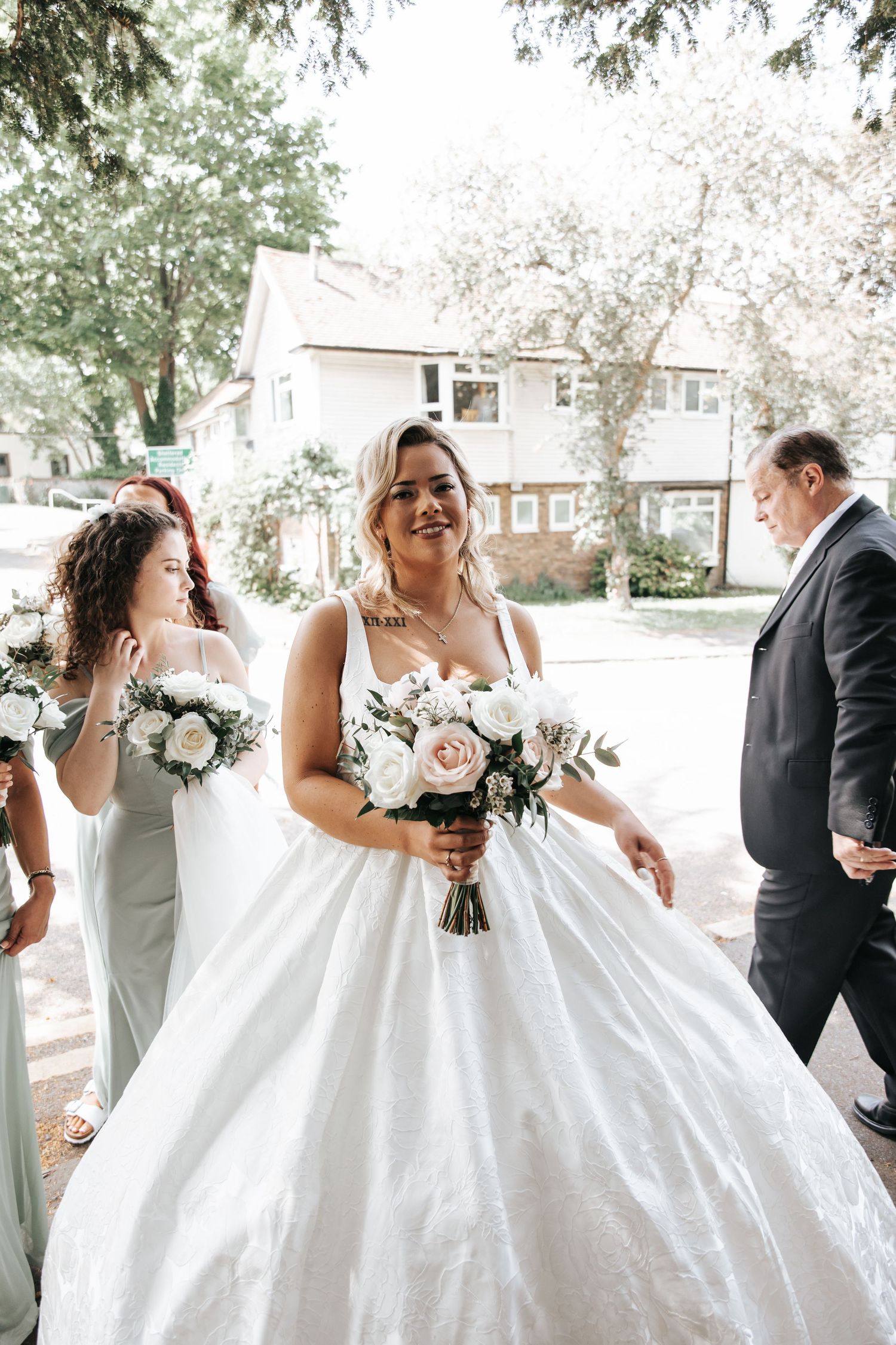 Bride in a white ball gown holds a bouquet of white and pink roses, smiling outdoors with bridesmaids nearby.