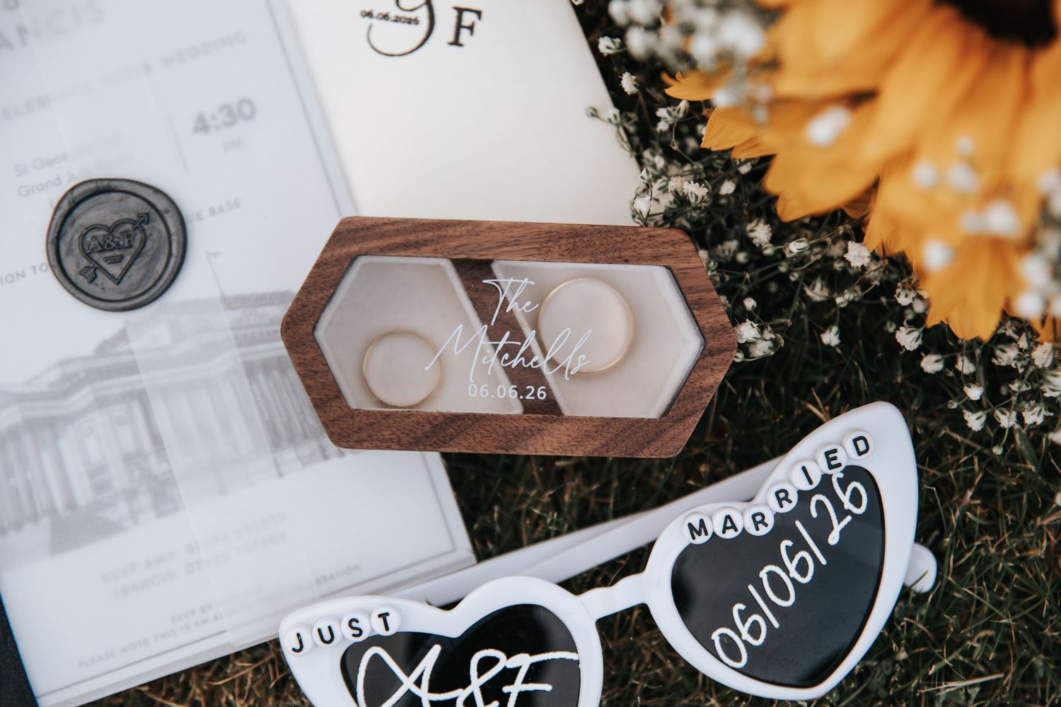 Wedding rings in wooden box with just married sunglasses and floral invitation suite on grass.