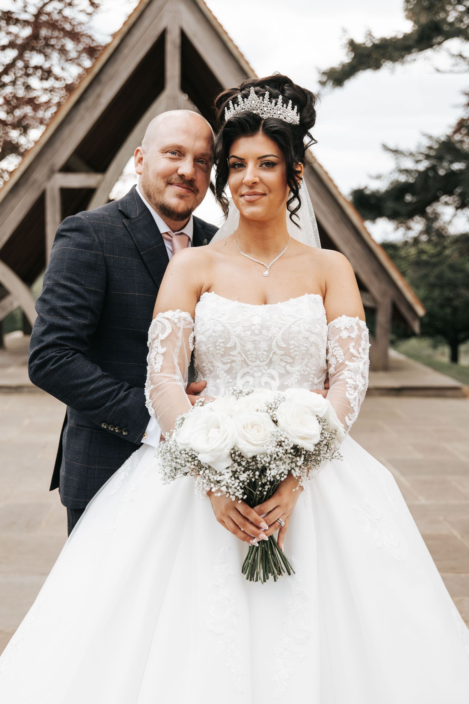 Bride in white ballgown with tiara holds rose bouquet beside suited groom outside wooden chapel.