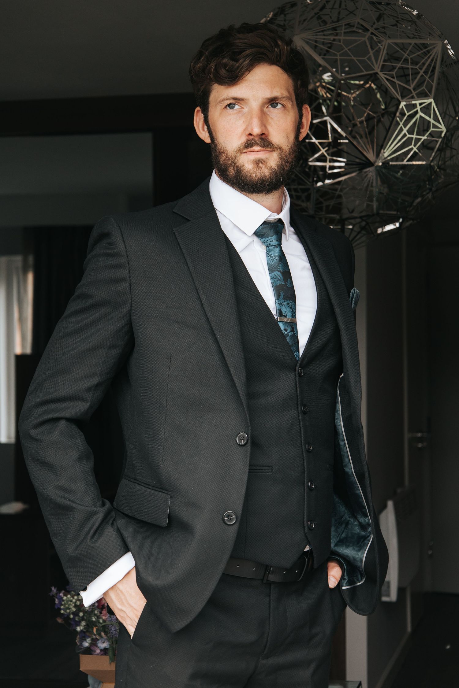 A man in a black suit and tie stands in a dimly lit interior space.