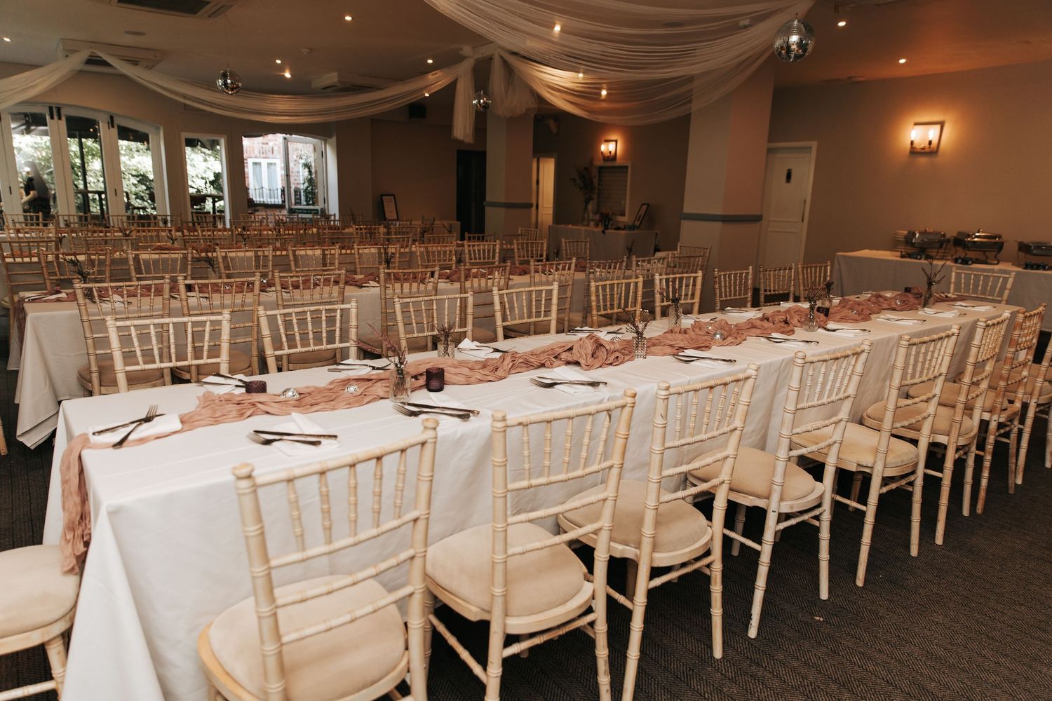 Elegant wedding reception hall with long white-clothed tables, chiavari chairs, and draped ceiling decor.