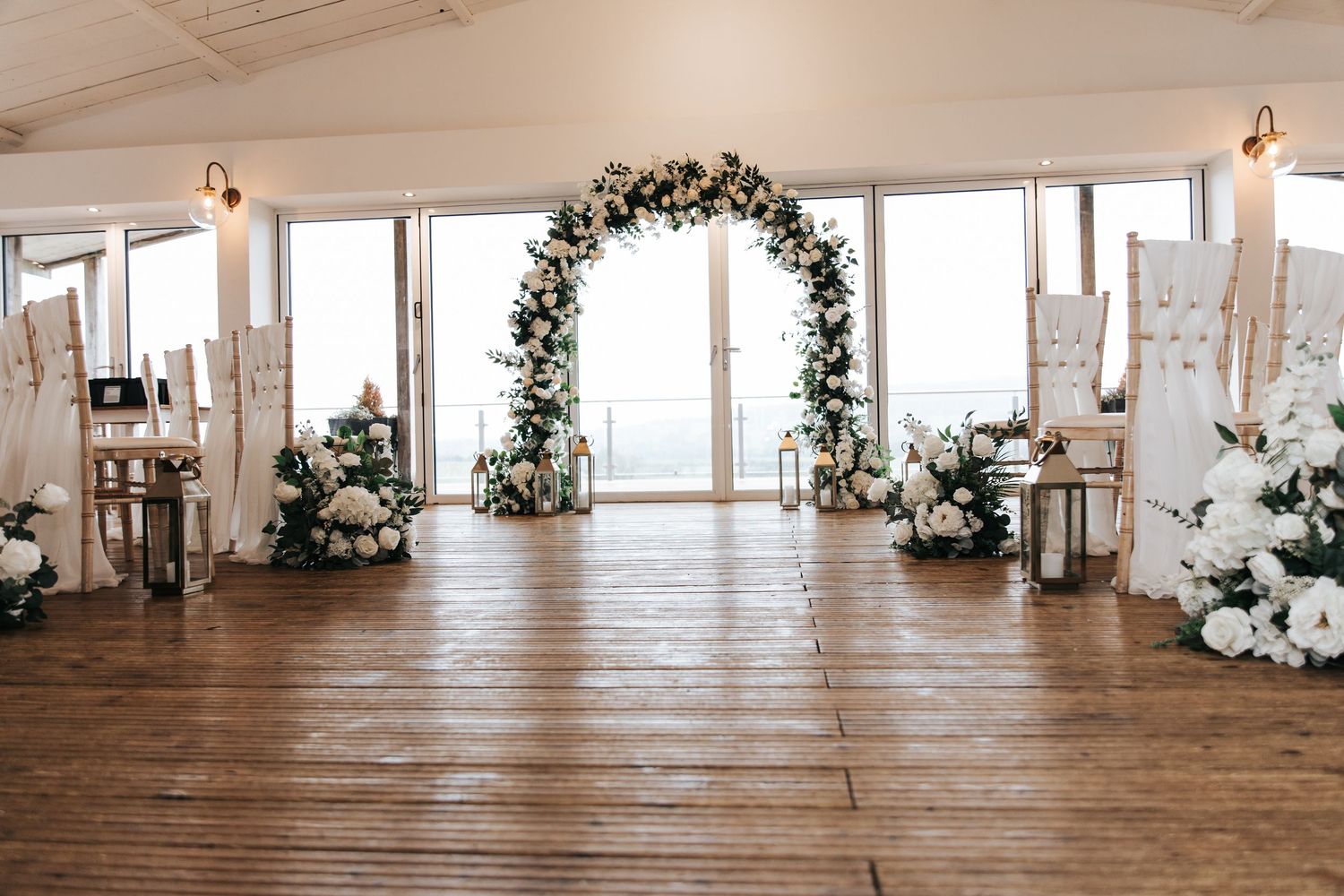 Elegant wedding ceremony venue with floral arch, white roses, wooden floor, and ocean view backdrop.