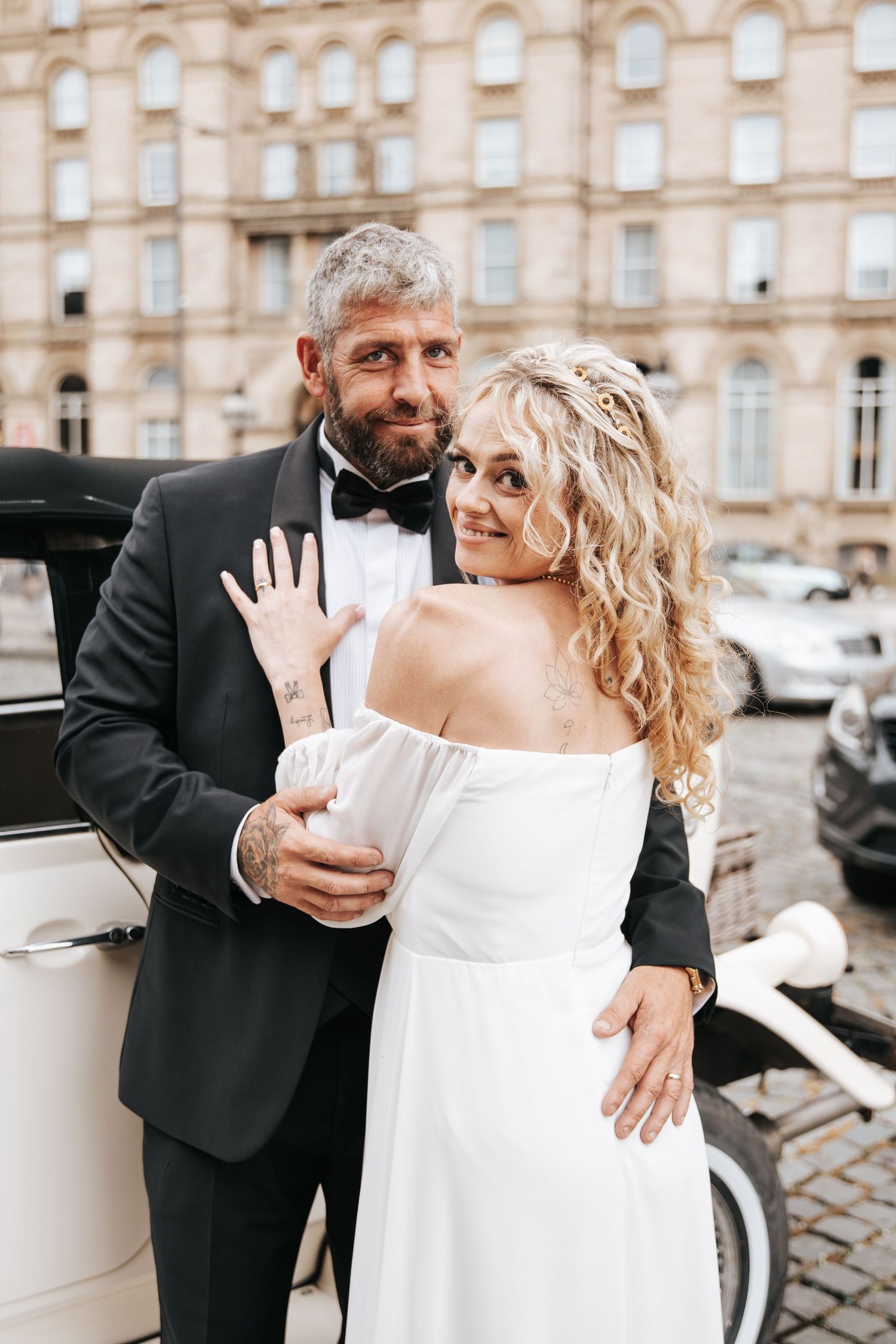Happy newlyweds pose by vintage white car, bride in off-shoulder gown showing ring, groom in black tuxedo with bow tie.