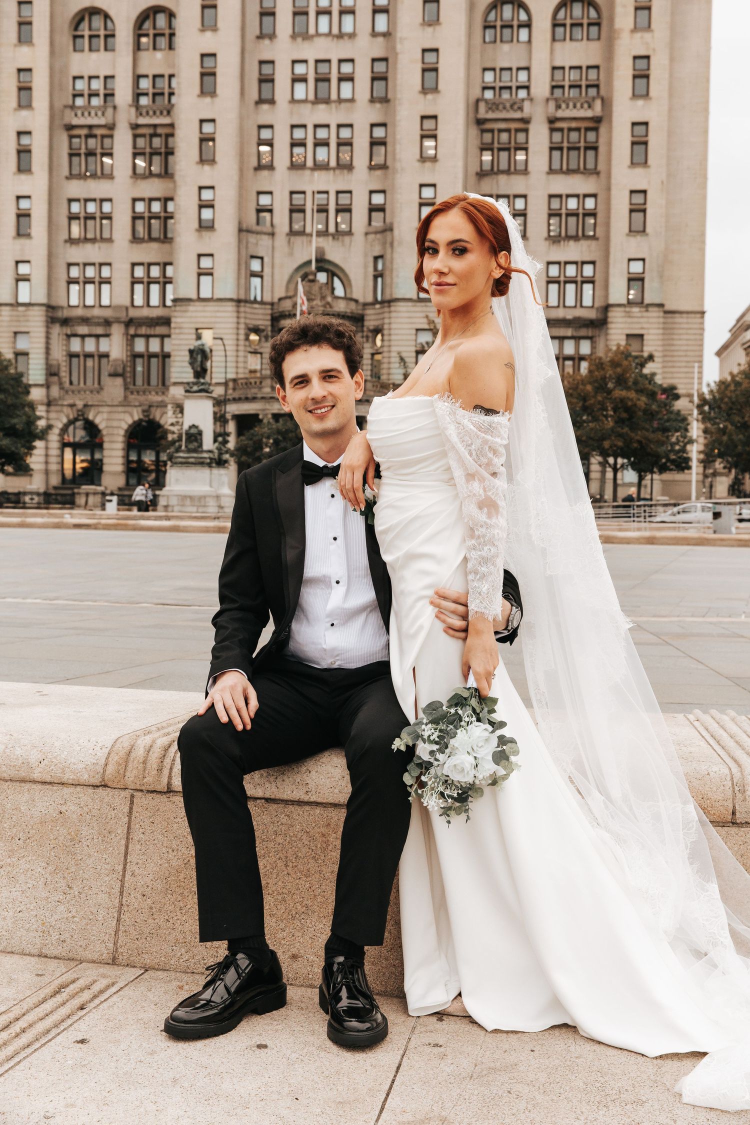 A bride in a white gown and groom in a black tuxedo pose together outdoors in front of a classic urban building.
