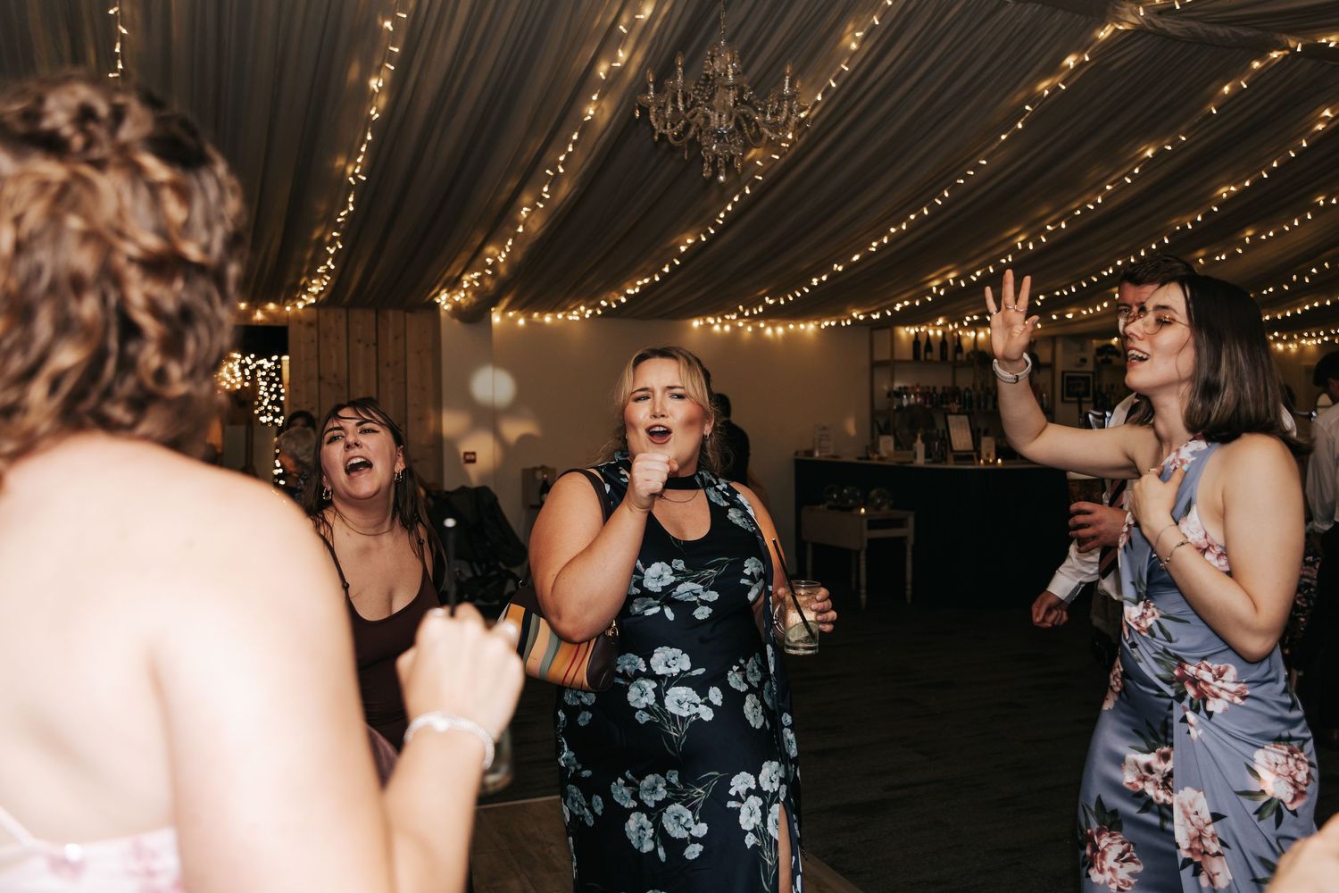 Women singing and dancing energetically at a wedding reception under fairy lights in a decorated venue.