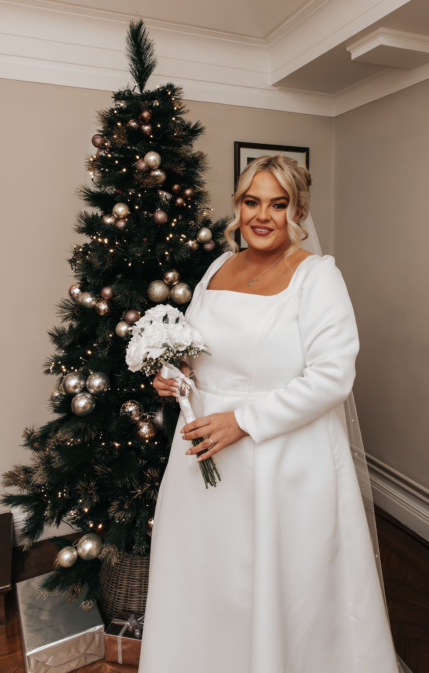 A winter wedding dress displayed near decorated Christmas trees with white long sleeves and elegant details.