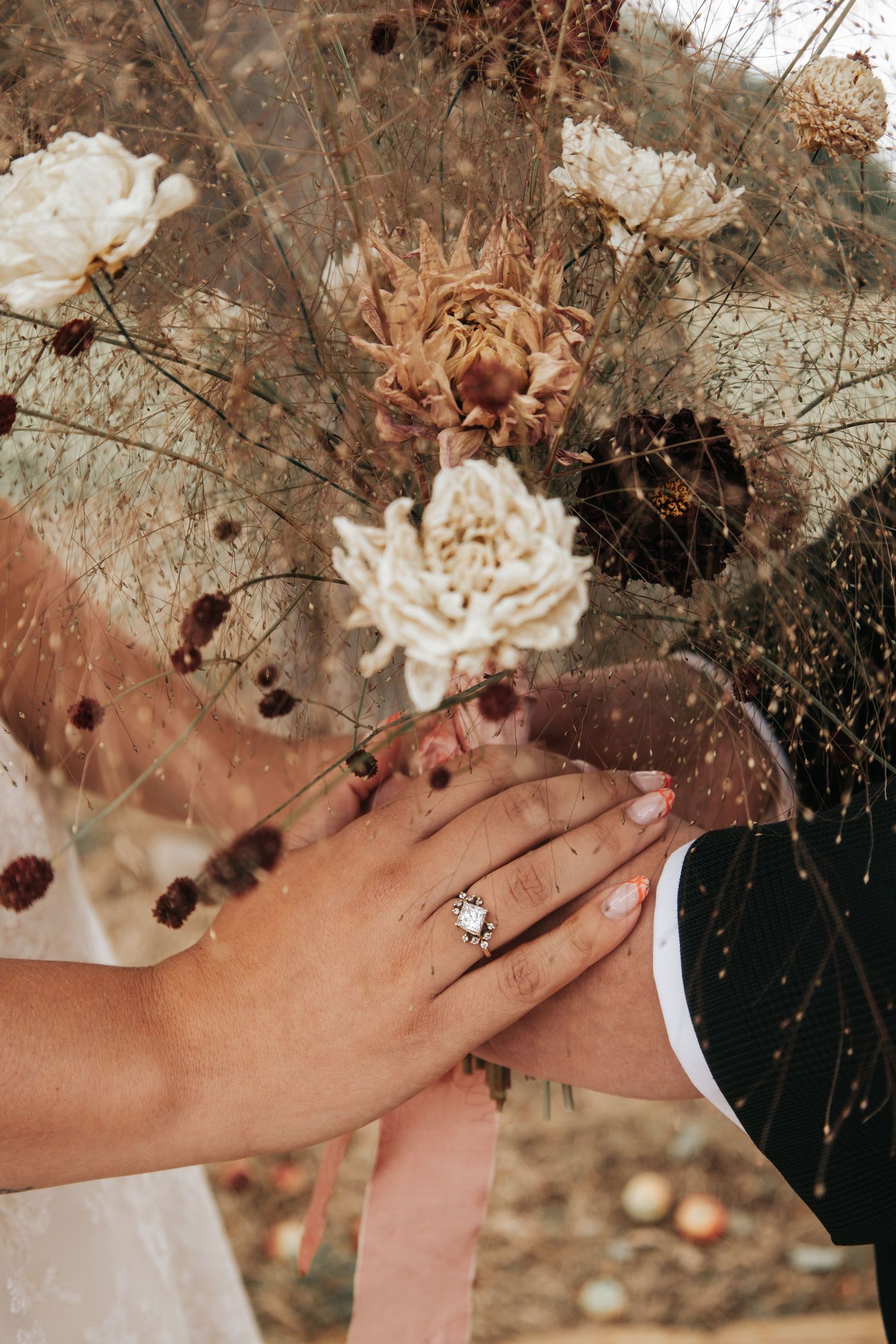 Close-up of hands displaying an engagement ring with delicate dried flowers in the background.