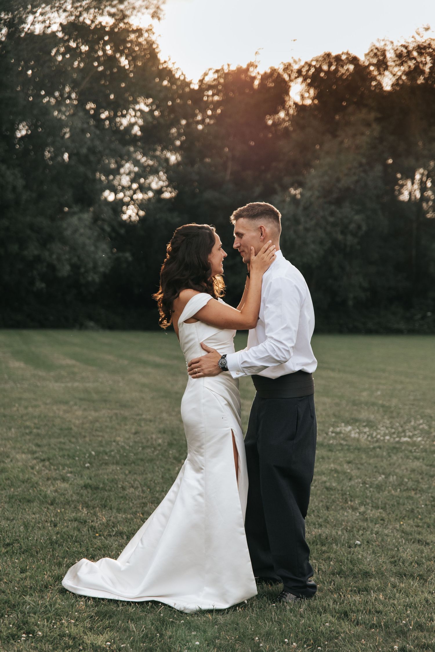 A bride and groom share a romantic embrace at golden hour on a lush green lawn surrounded by trees.