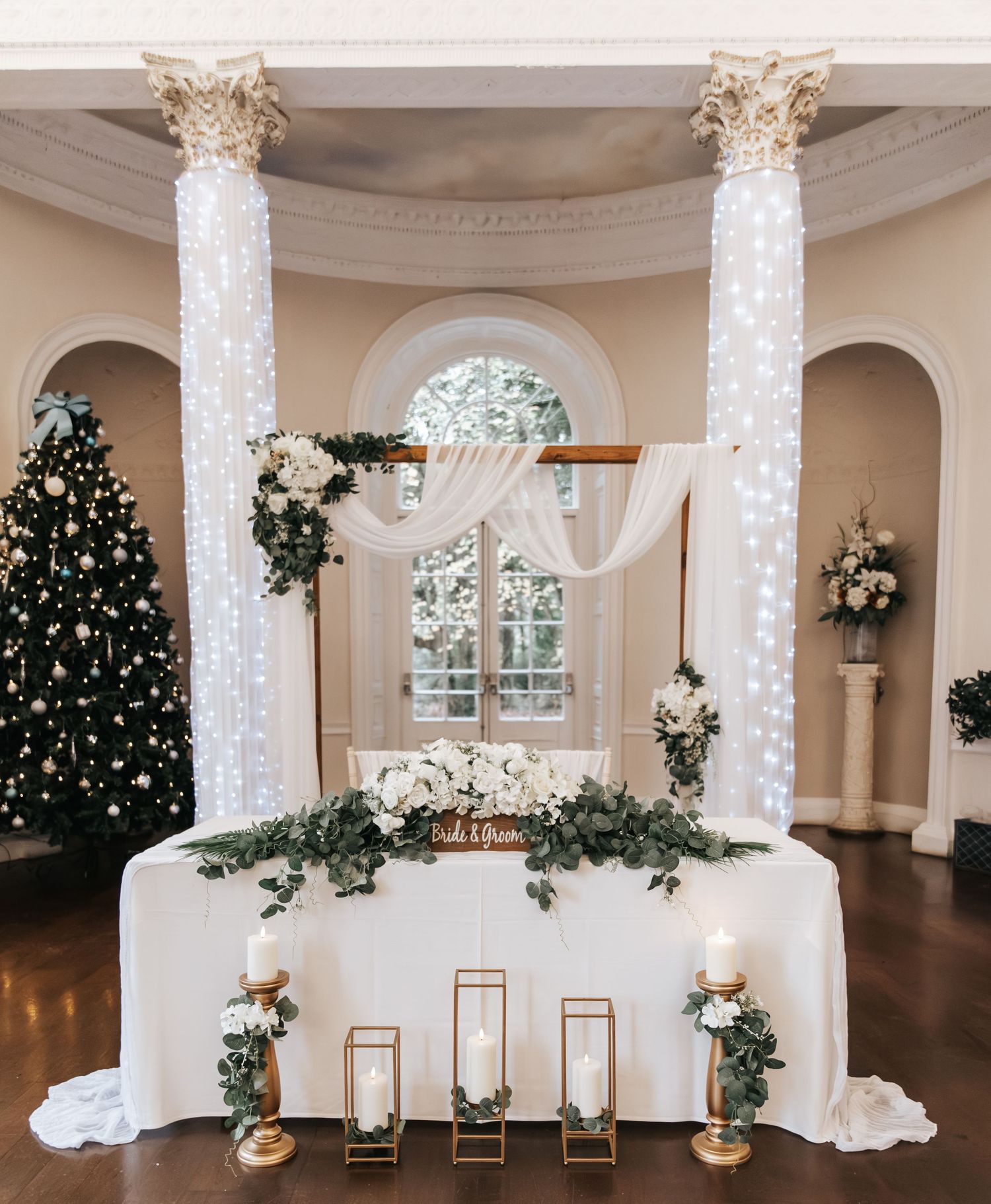 Elegant wedding sweetheart table with white draping, greenery garland, gold candle holders, and fairy light columns in a ballroom.