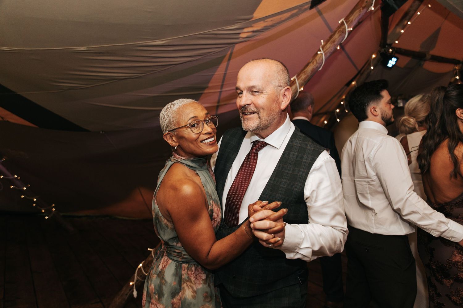 Couple dancing joyfully at a wedding reception inside a decorated tent with fairy lights.