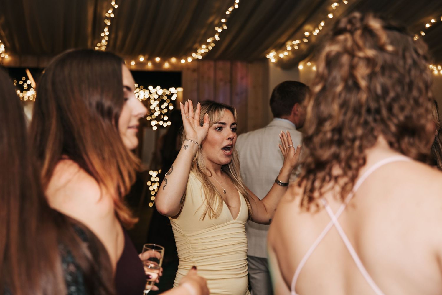 Group of women dancing and laughing at a wedding party with warm fairy lights overhead.