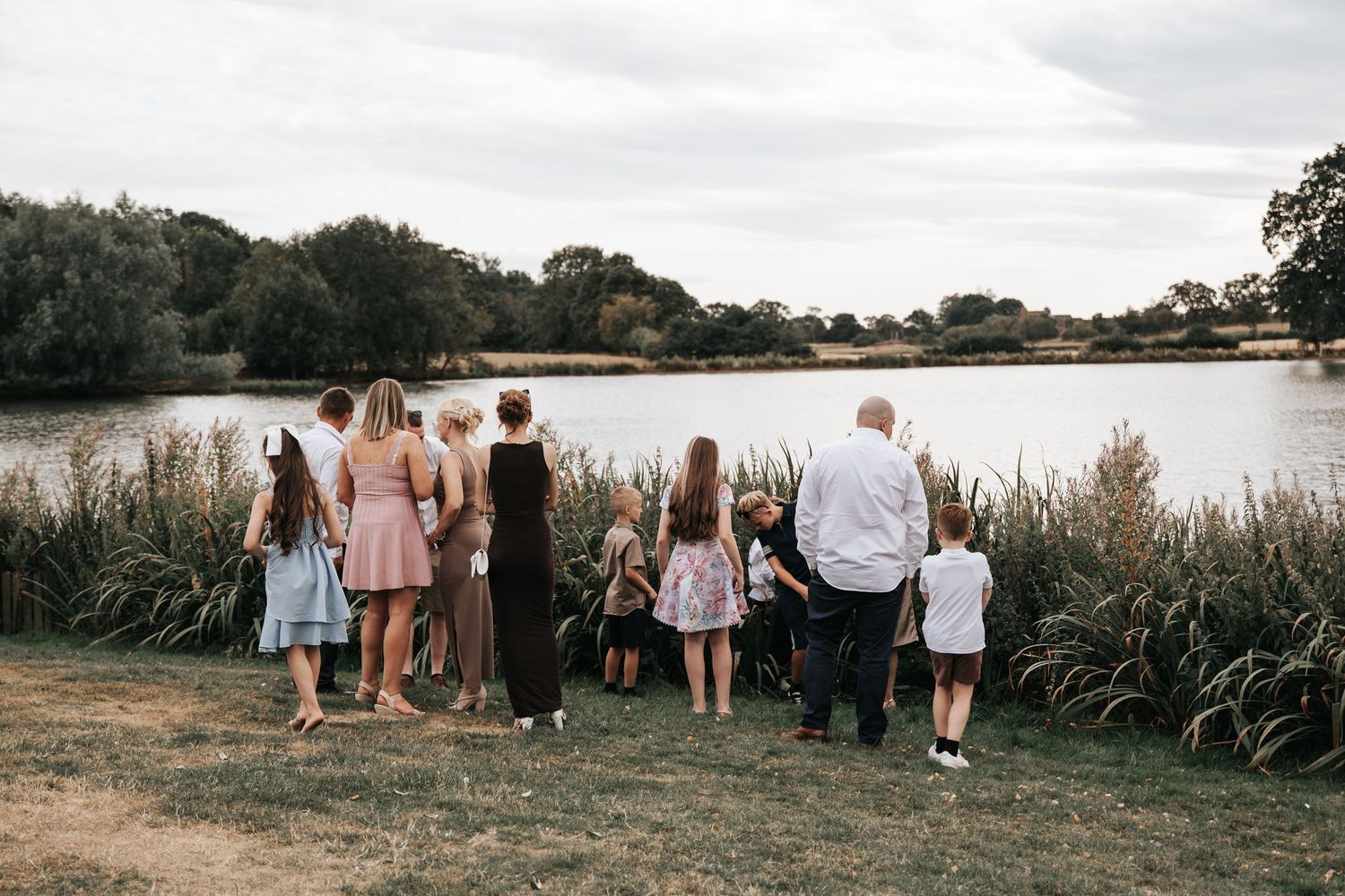 Large family group standing together by a serene lake surrounded by tall grass and trees on a cloudy day.