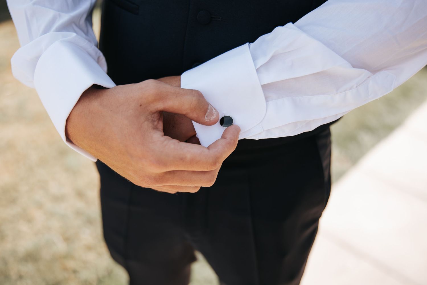 Man in white dress shirt and black vest fastening a cufflink on his sleeve.