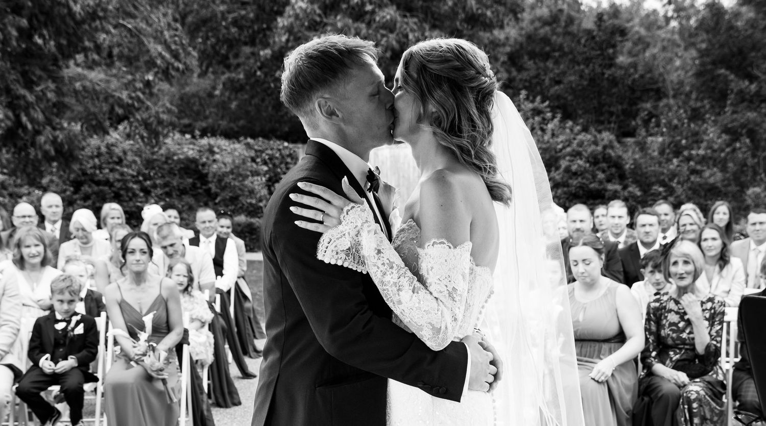 Black and white wedding photo of bride and groom sharing first kiss at outdoor ceremony with guests.