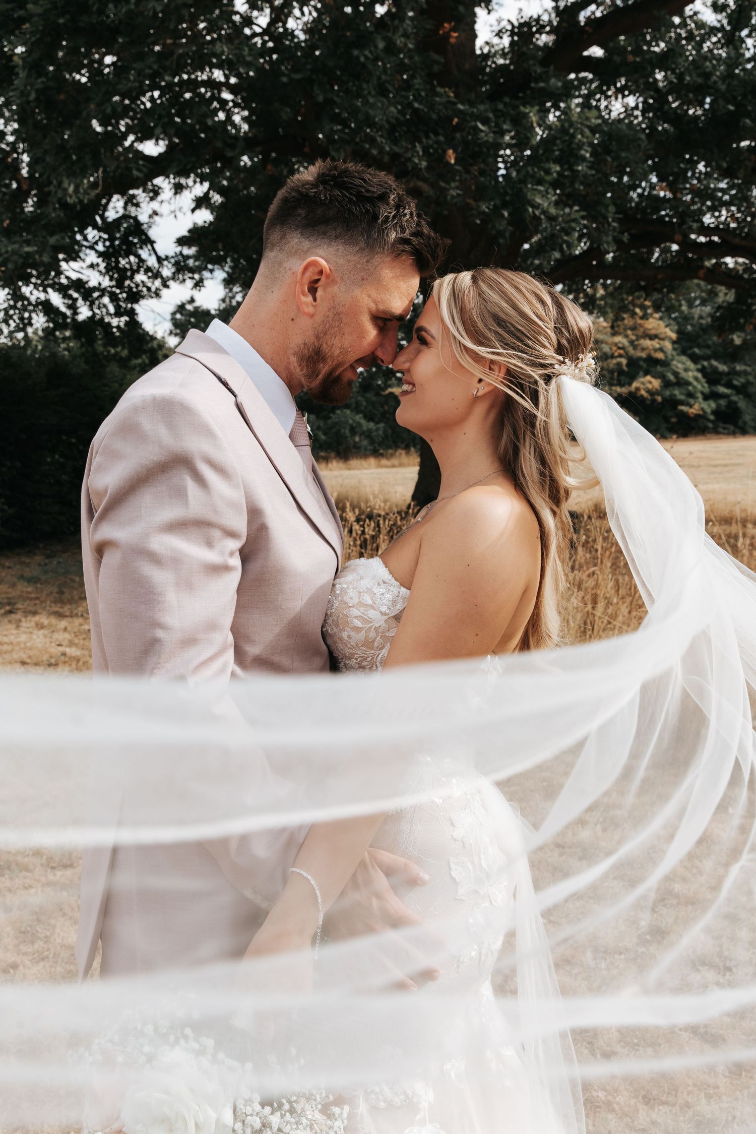 A bride and groom share a romantic moment outdoors, her flowing white veil billowing around them near a grand tree.