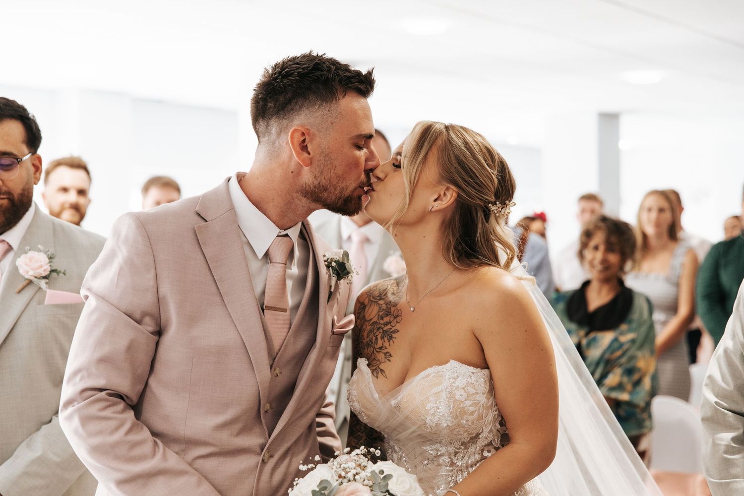 Bride and groom share first kiss at wedding ceremony, groom in blush suit, bride in lace strapless gown holding bouquet.