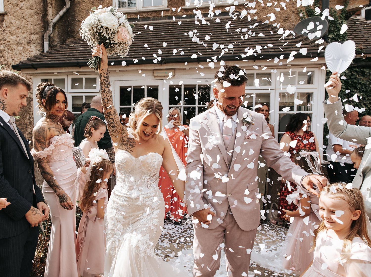 Bride and groom walk through confetti shower with wedding guests cheering outside a rustic venue.