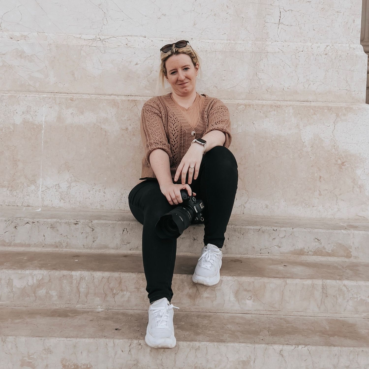 Woman sitting on stone steps holding a camera, wearing black jeans and white sneakers, smiling outdoors.