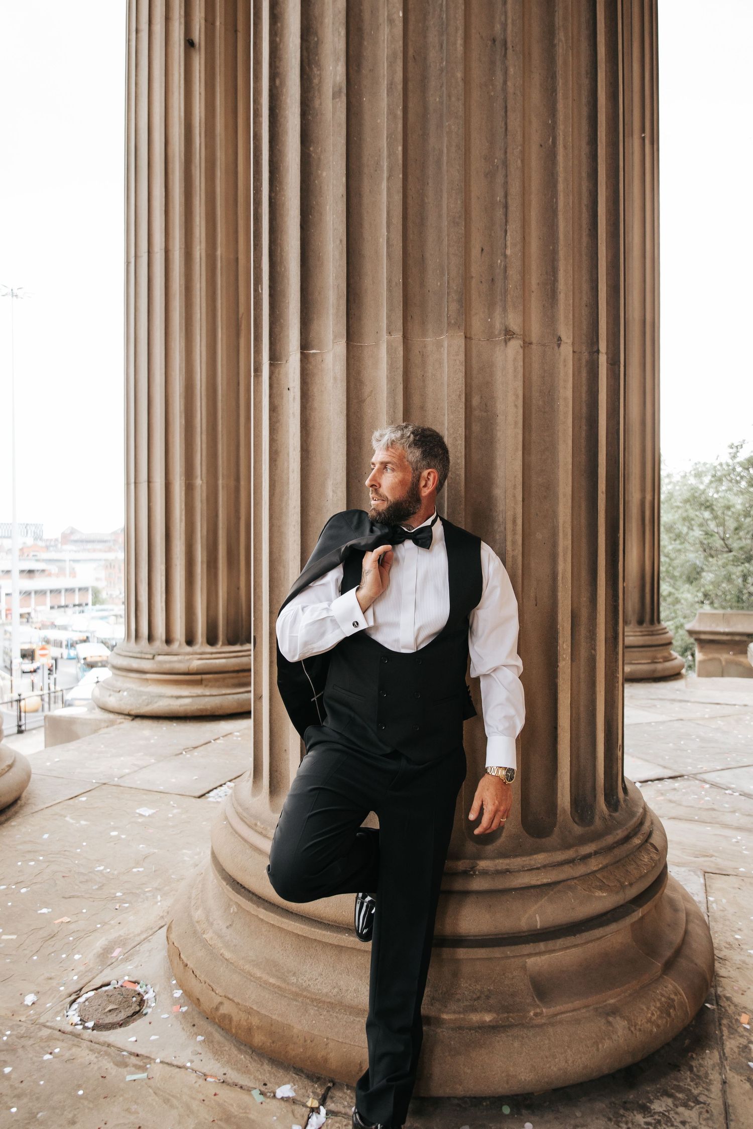 A dapper man in a black vest and bow tie leans against a grand stone column outdoors.