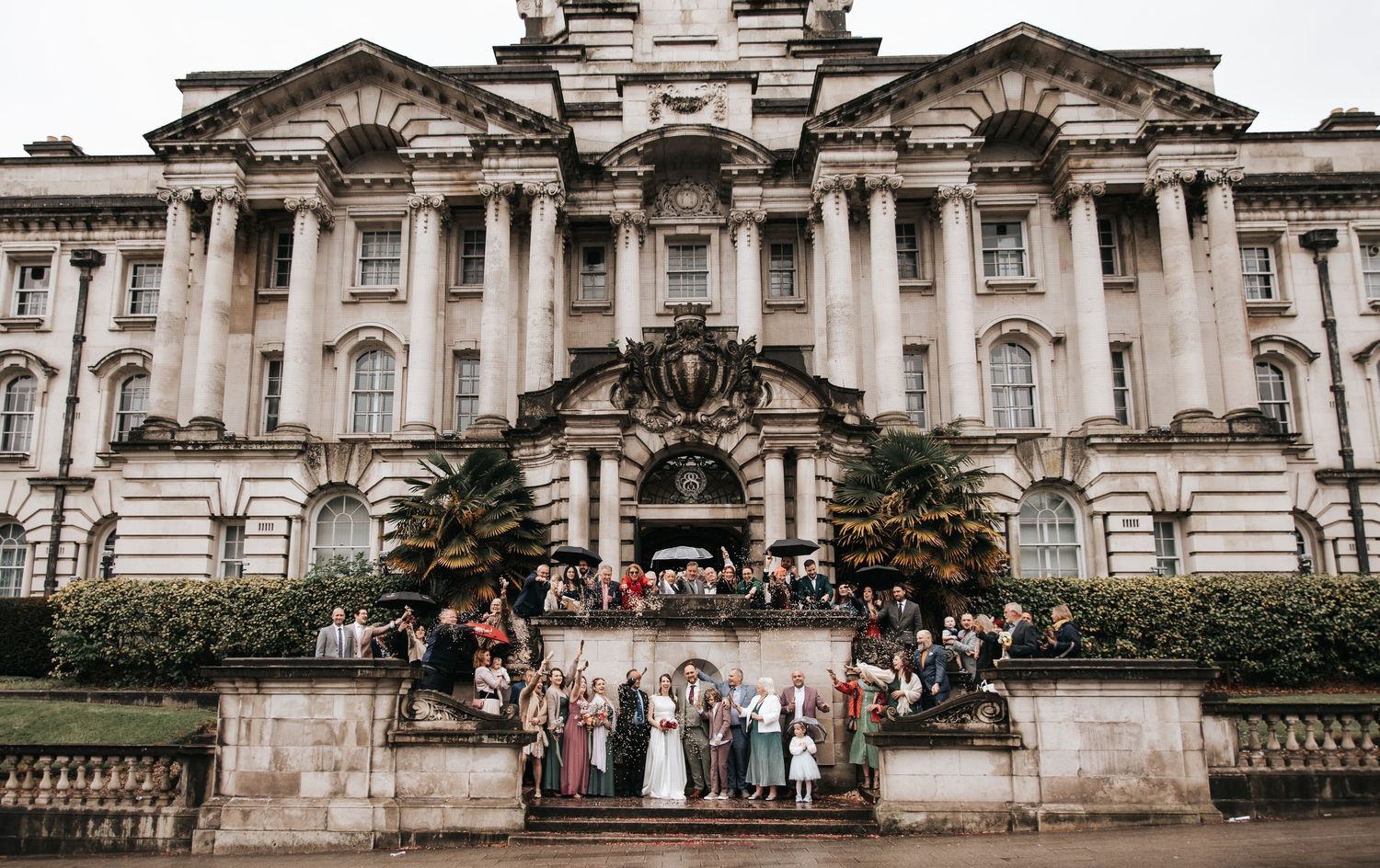 Wedding party posing in front of a grand neoclassical building with ornate facade and lush greenery.