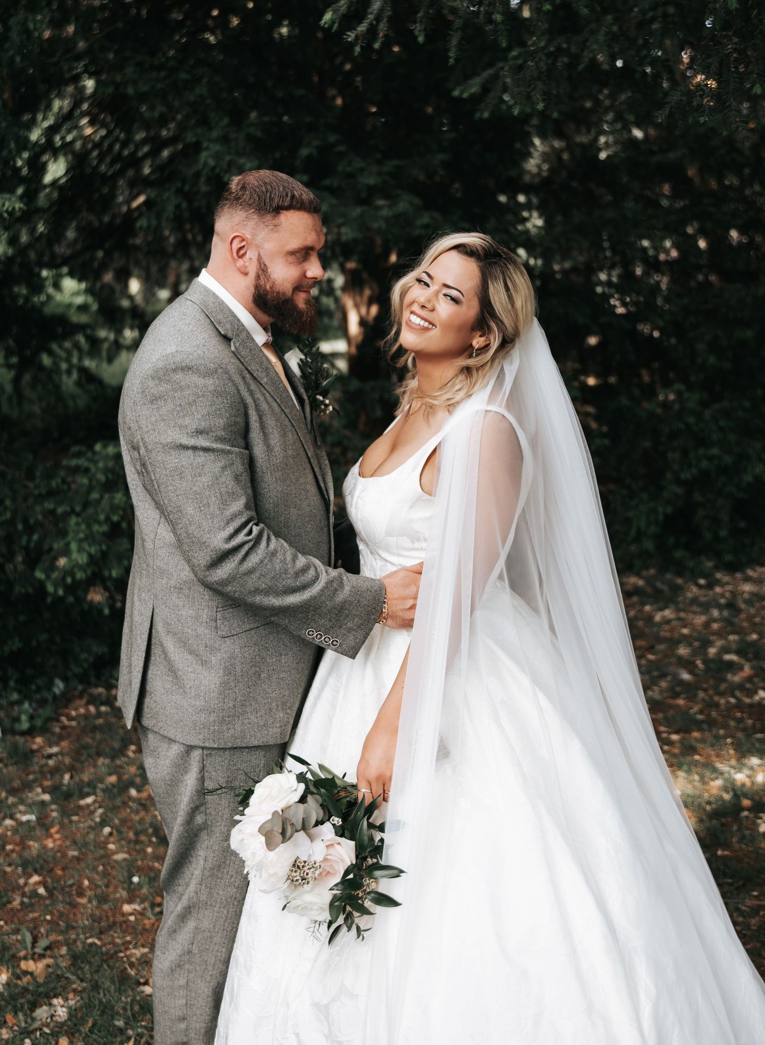Bride in white gown with veil holding bouquet smiles at groom in gray suit outdoors.