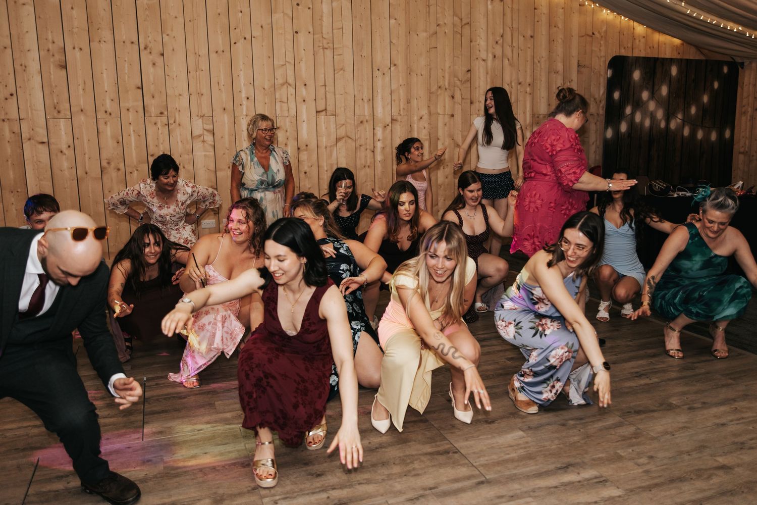 Guests dancing and having fun at a lively wedding reception on a wooden dance floor.
