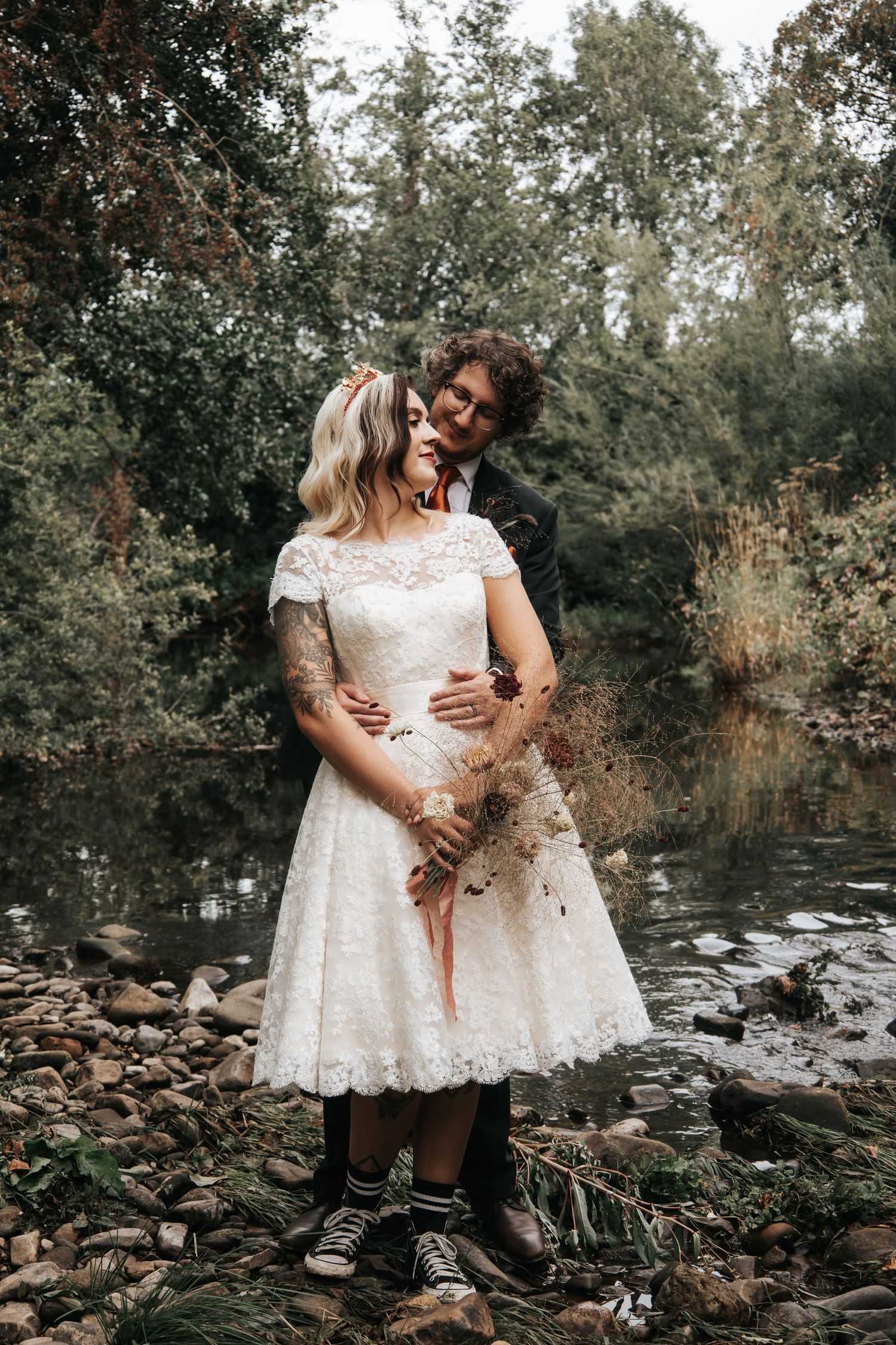 A romantic maternity photoshoot by a rocky stream in a wooded setting features a white lace dress.