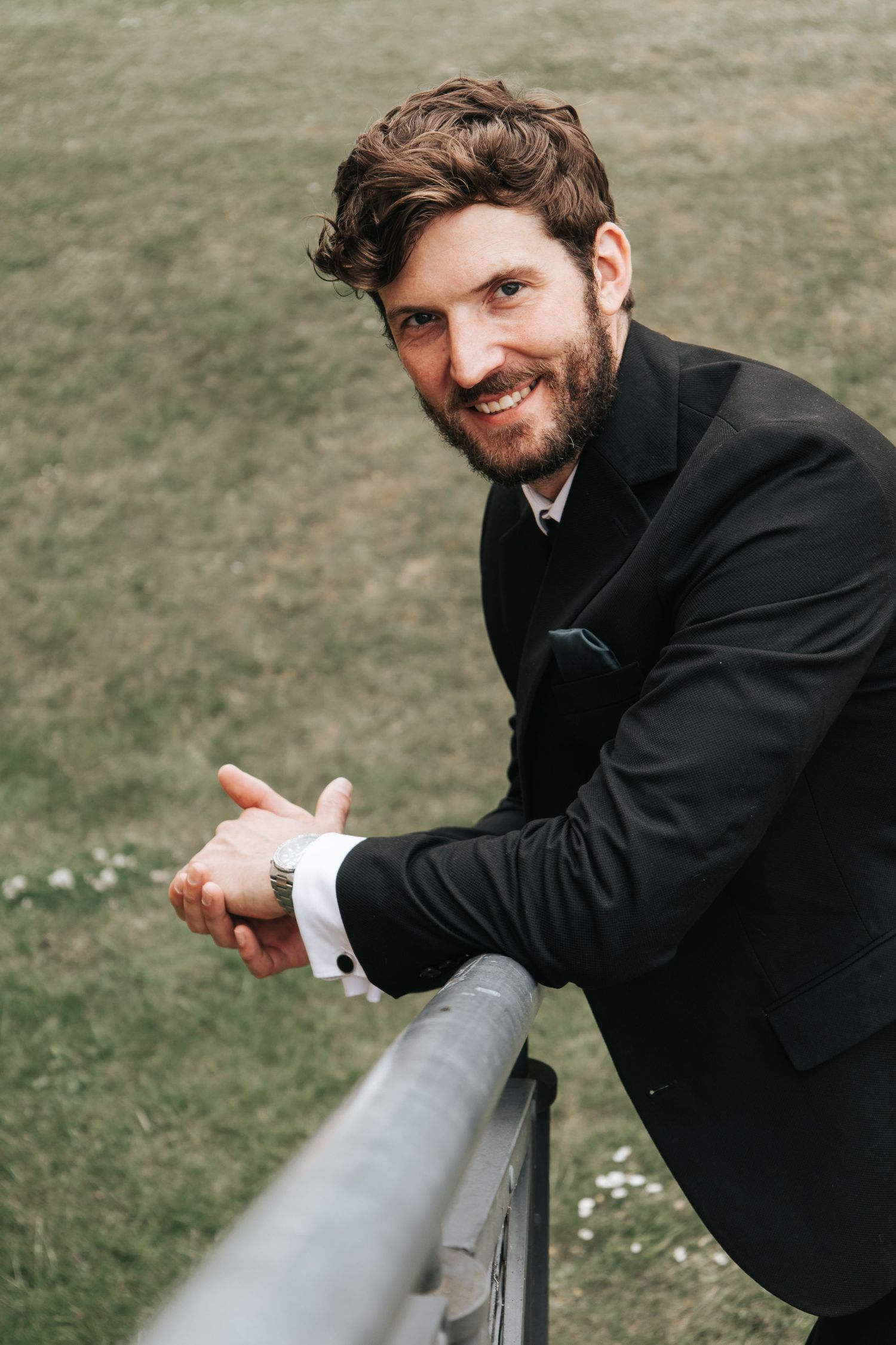 Groom in black suit leaning playfully on outdoor railing.