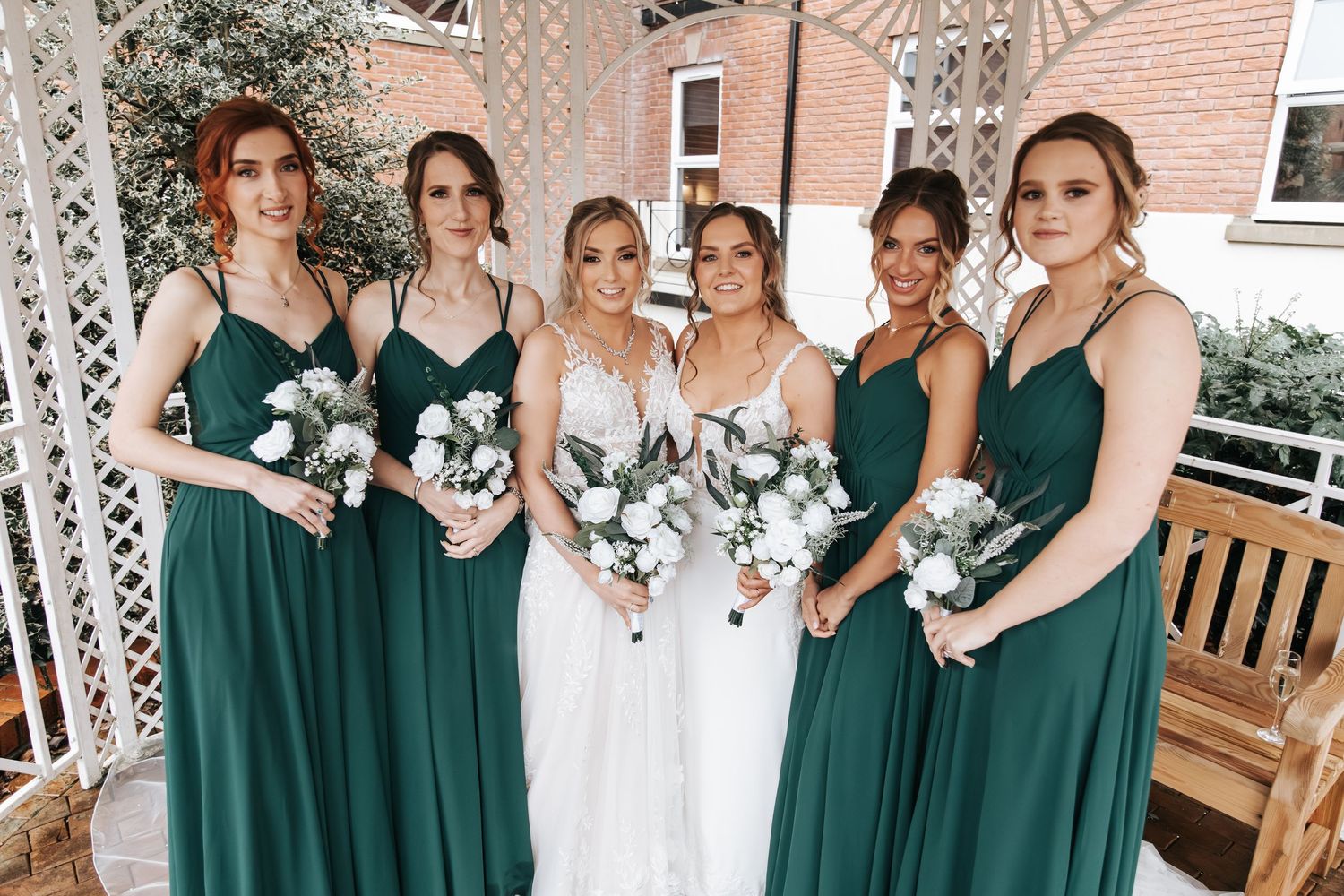 Wedding party in emerald green dresses and white gowns pose in outdoor setting.