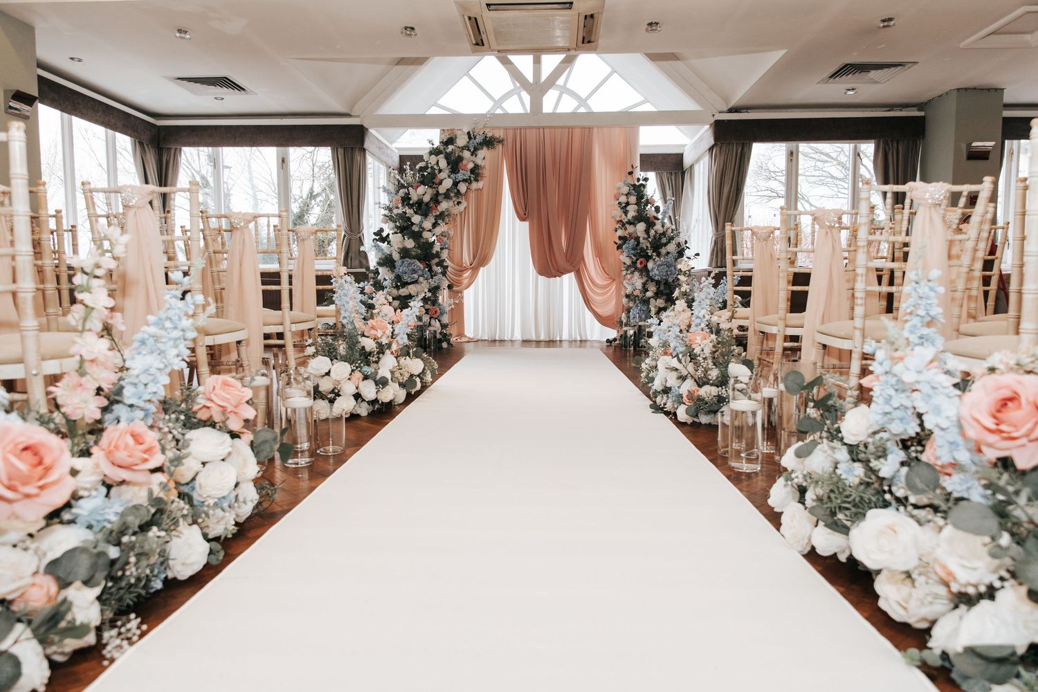 Elegant wedding aisle with white runner, pink and white floral arrangements, gold chairs, and draped blush curtains.