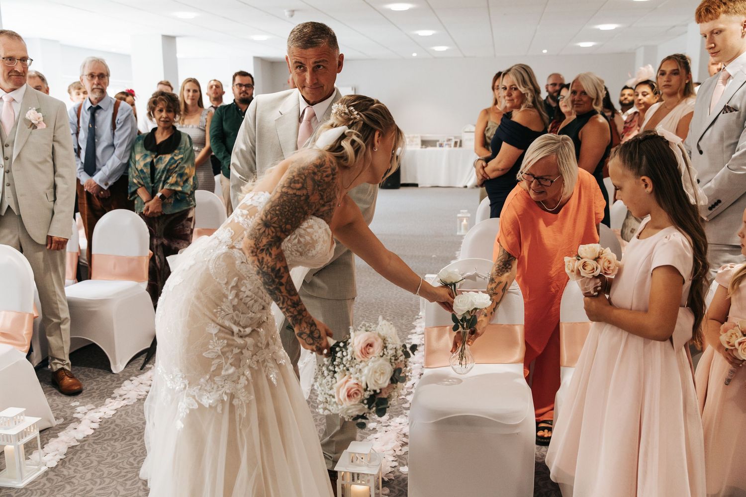 Bride in lace wedding dress with tattoos greets flower girl during indoor wedding ceremony with guests seated in background.