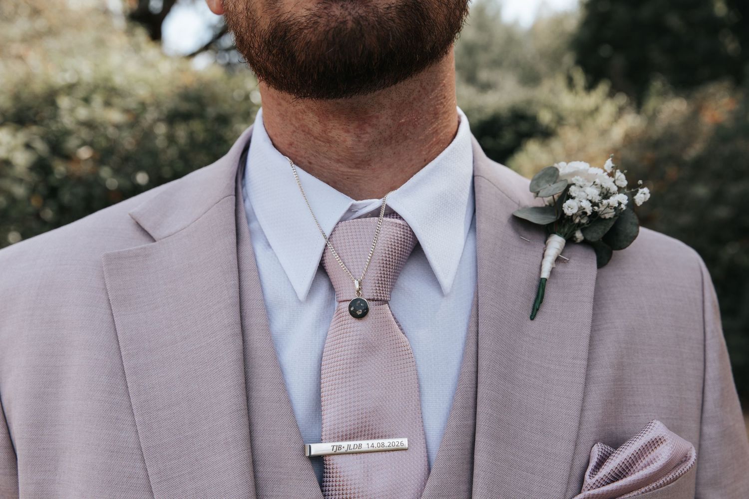 Groom in blush pink suit with matching tie, silver tie bar, boutonniere, and pocket square at wedding.