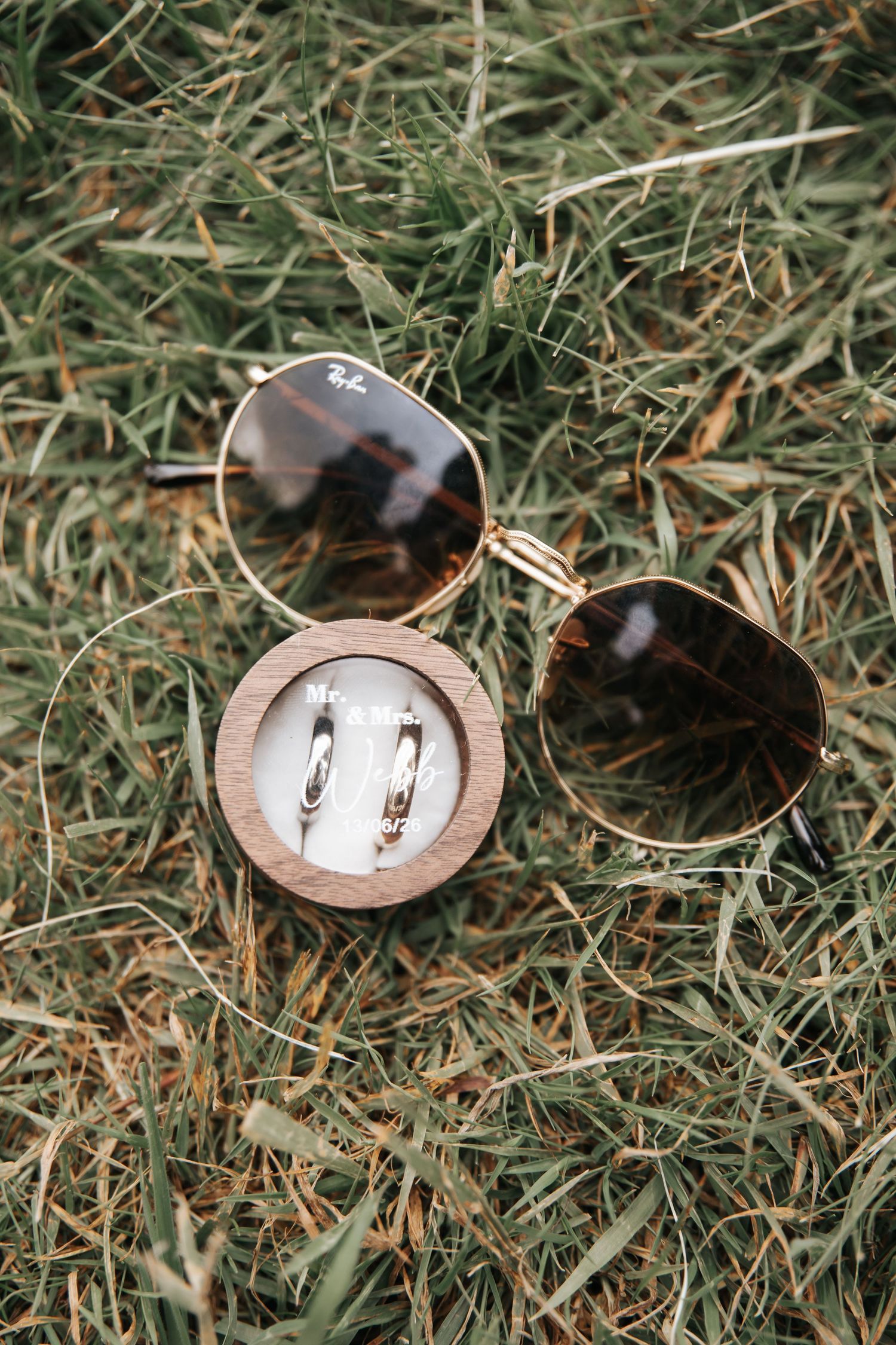 Ray-Ban sunglasses and wedding rings in a wooden box on green grass.