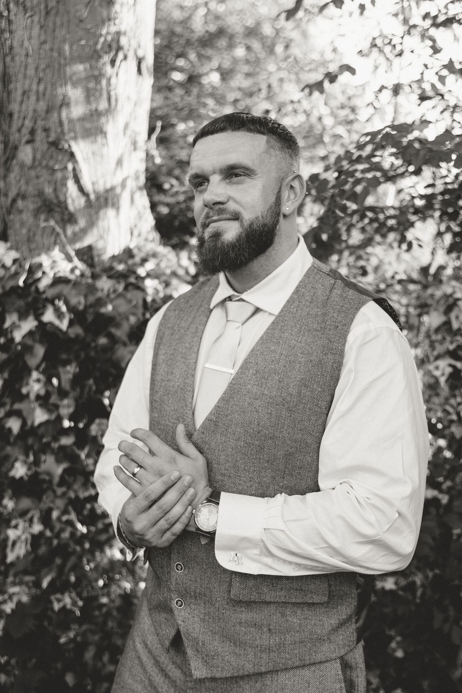 Black and white portrait of a bearded groom in a tweed vest and tie, standing outdoors among lush foliage.