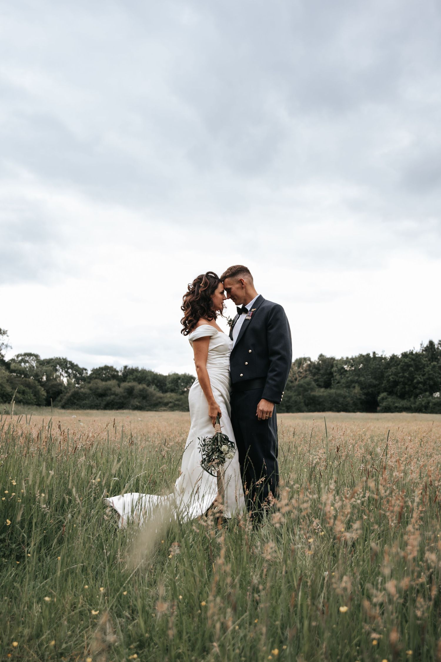 Bride and groom embrace in a lush meadow, she in a white gown holding a bouquet, he in a dark suit, under cloudy skies.