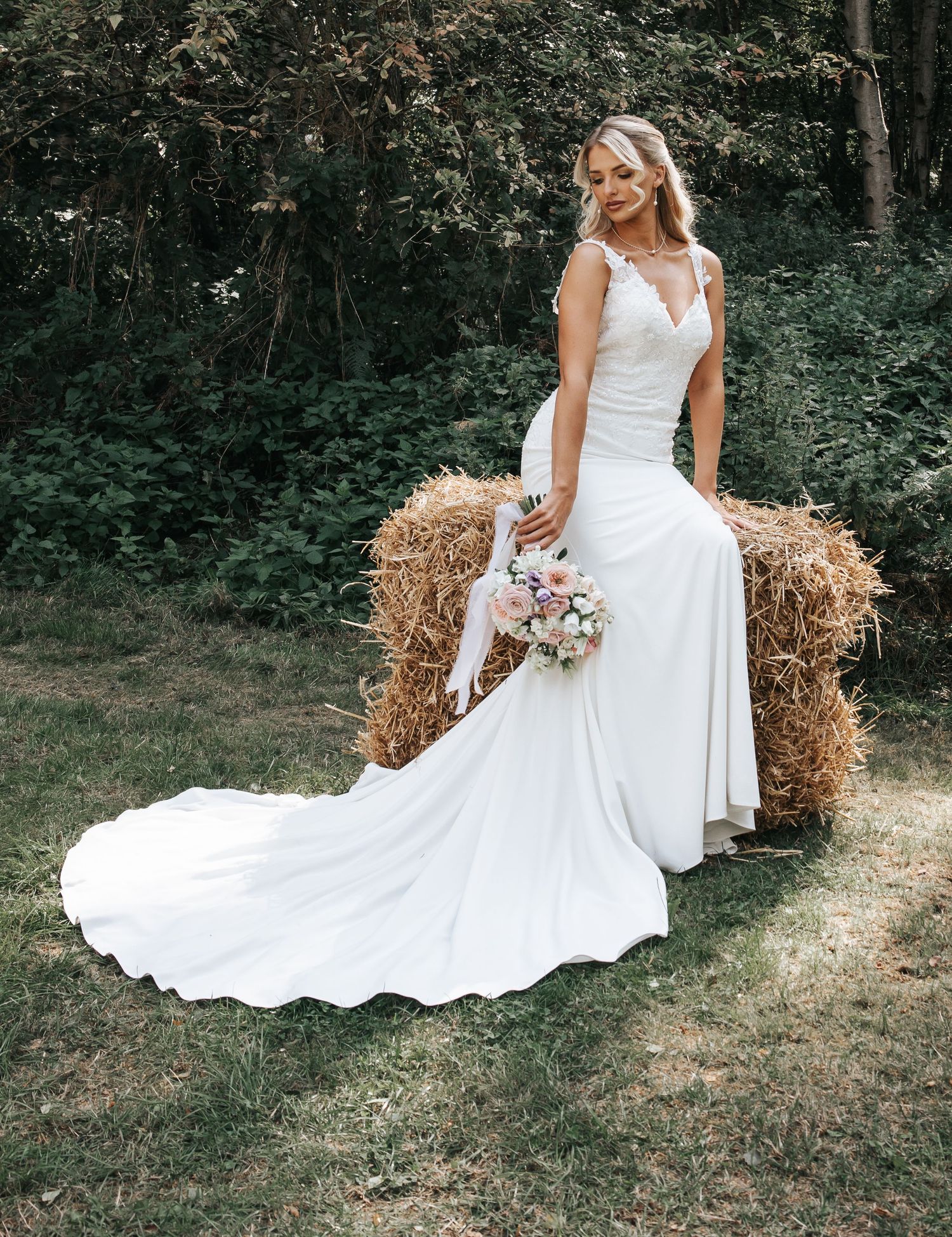 Full-length view of flowing white wedding dress as bride poses by hay bales in forest setting.