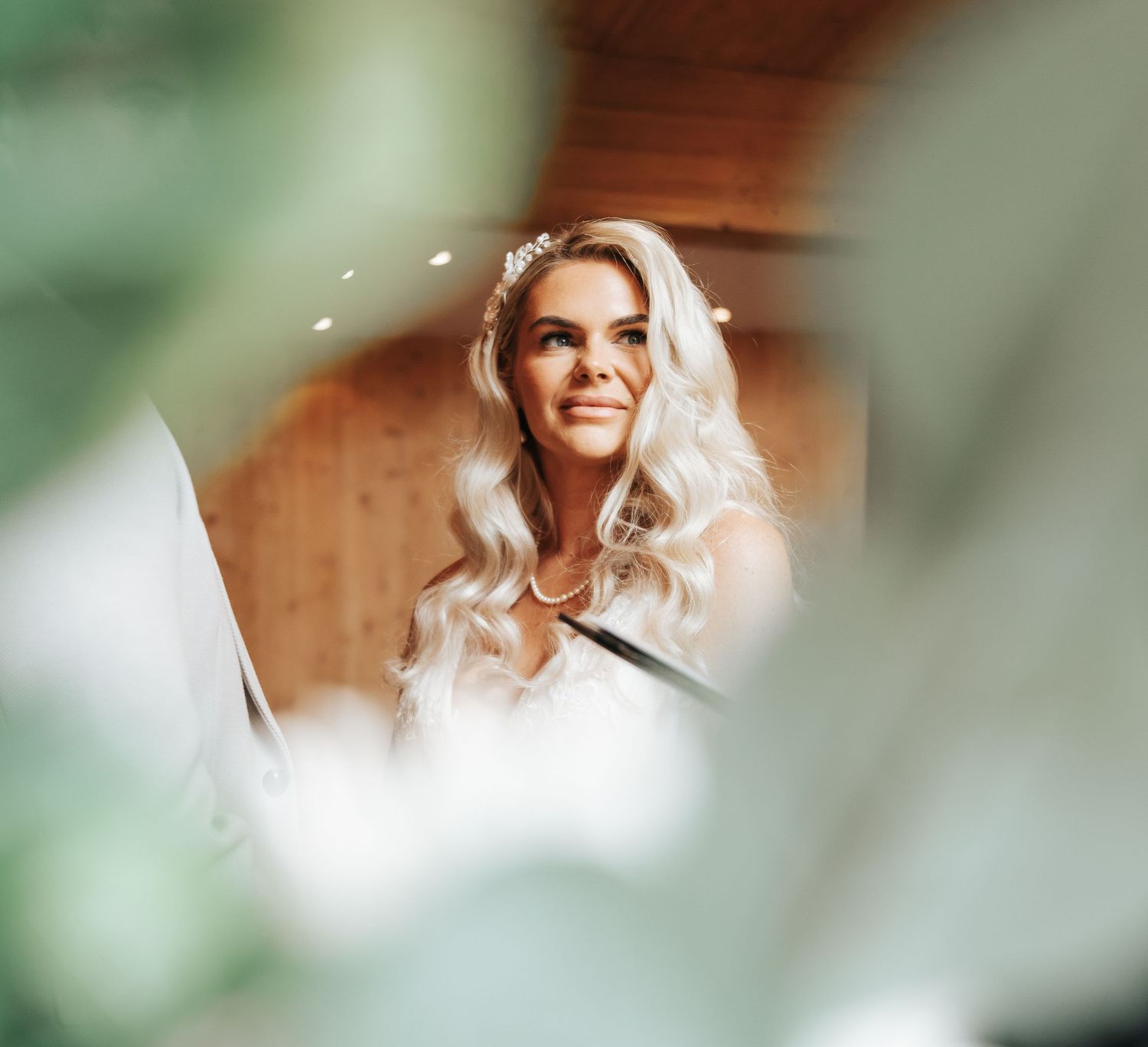 Smiling blonde bride with floral hair accessories wearing a white dress, photographed through green leaves in a wooden venue.