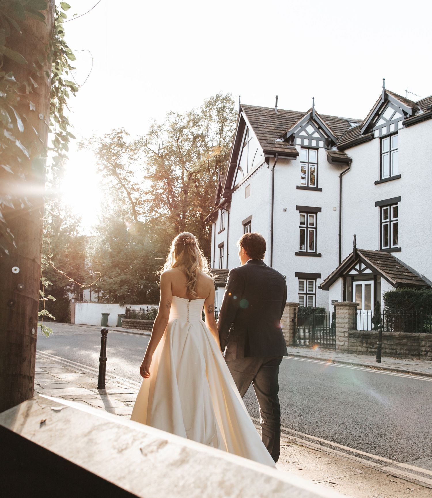A bride in a white gown and groom in a suit walk toward a sunlit street with a white Tudor-style building.