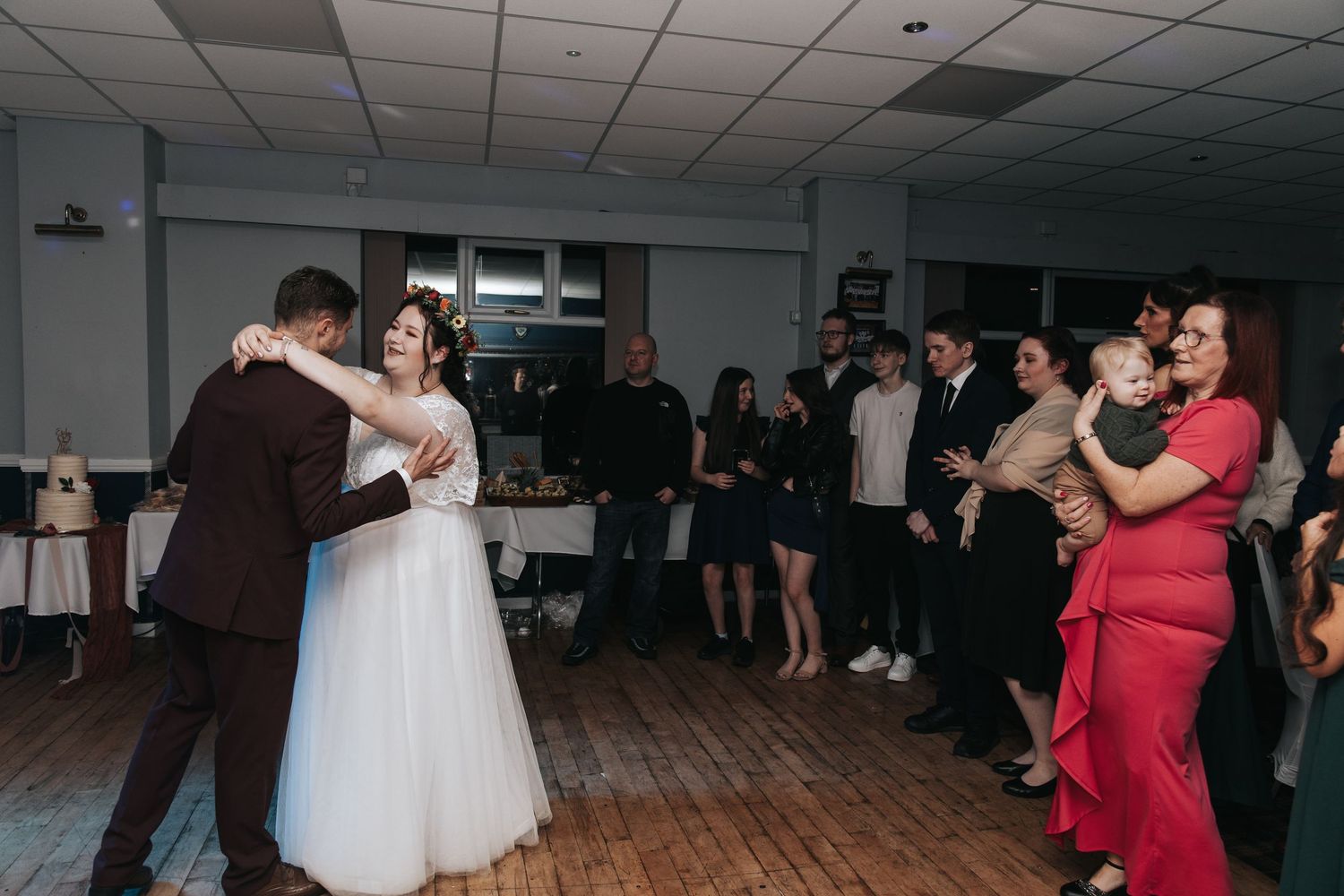 A group of people dancing at a wedding reception in a banquet hall with white ceiling tiles and wood flooring.