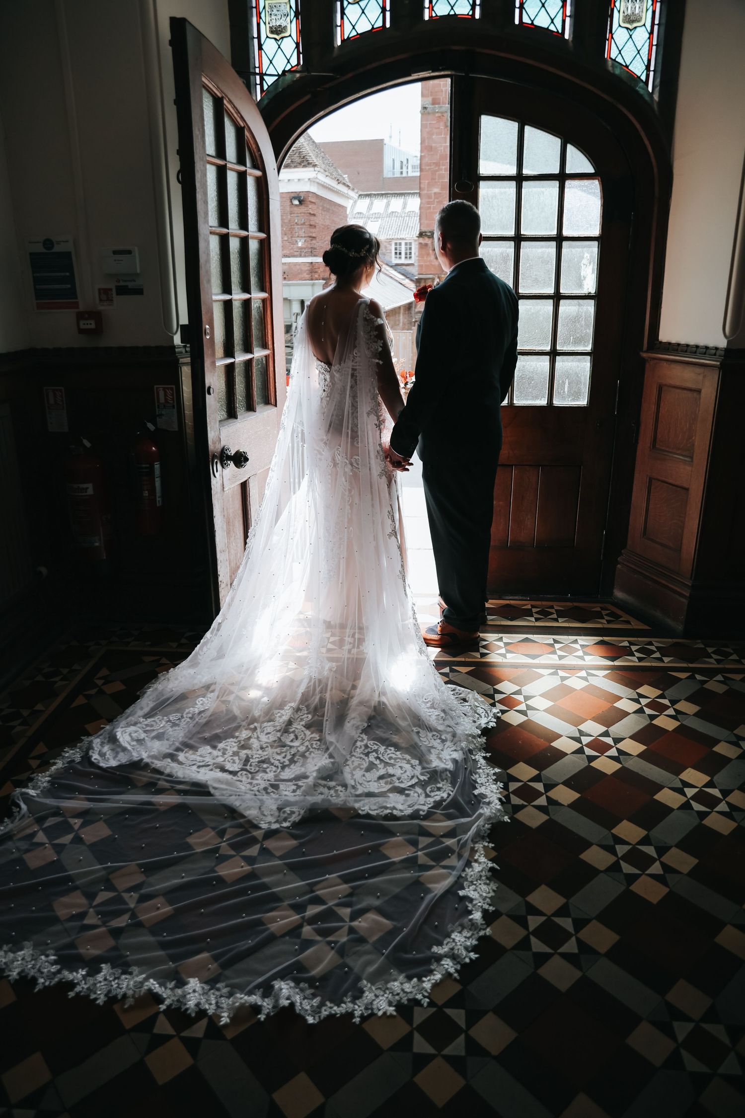 Bride and groom holding hands at church doors, her lace cathedral veil trailing on tiled floor in soft backlight.