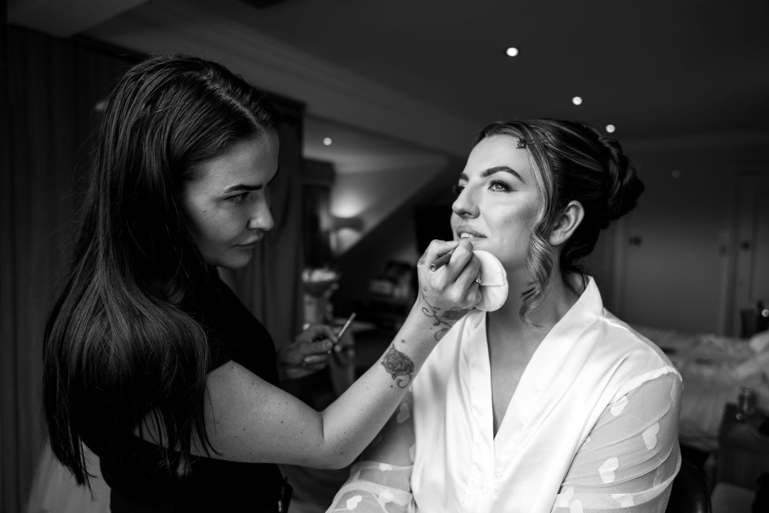 Black and white photo of a makeup artist applying lipstick to a bride in a white robe before her wedding.
