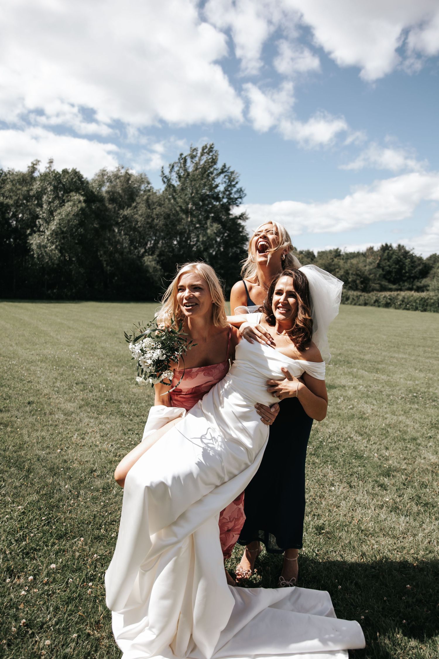 Bride being lifted by two laughing bridesmaids in colorful dresses on a sunny green lawn.