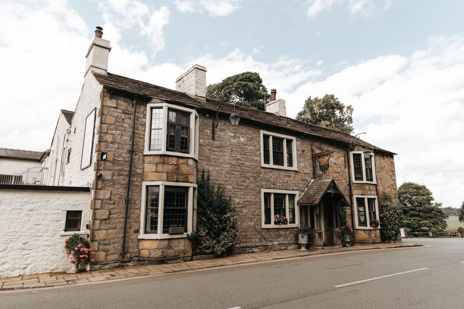 A charming stone-built country inn with mullioned windows and a covered entrance, set along a quiet village road under a cloudy sky.