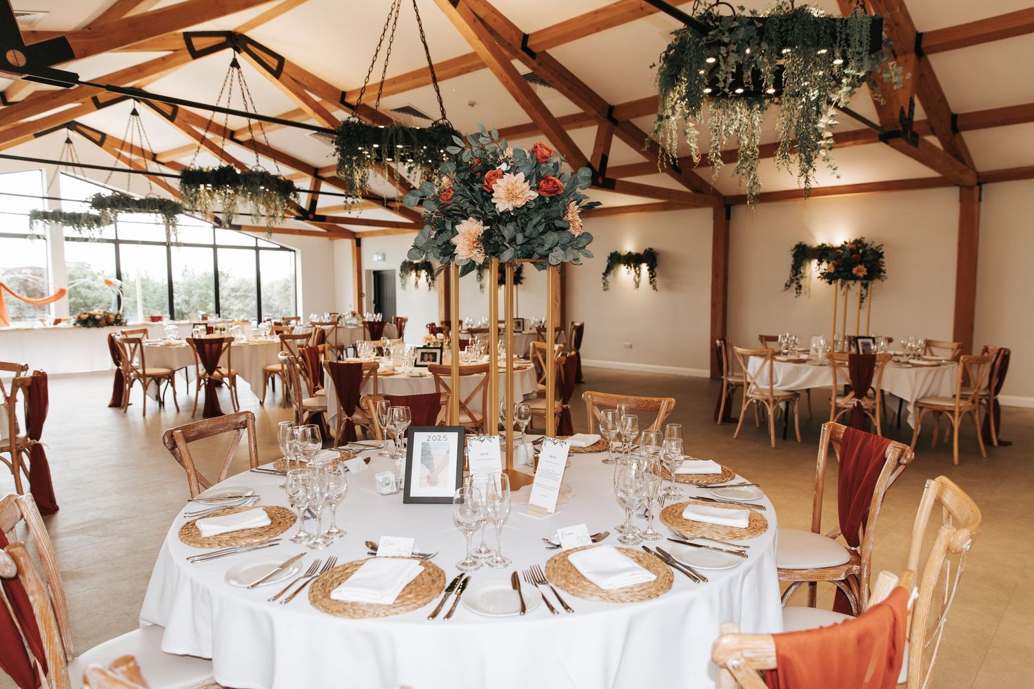 Elegant wedding reception hall with round tables, white linens, gold centerpieces, and lush floral ceiling installations.